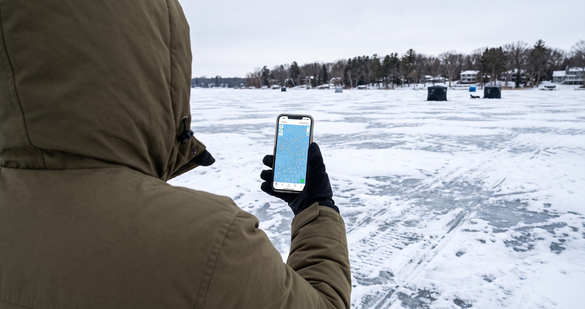 Over-the-shoulder view of an angler in an olive hooded parka holding up a smartphone running the Fish Guide Pro contour map app, with a snow-covered frozen lake, several dark ice fishing shelters, snowmobile tracks, and a tree-lined shoreline with homes in the background.
