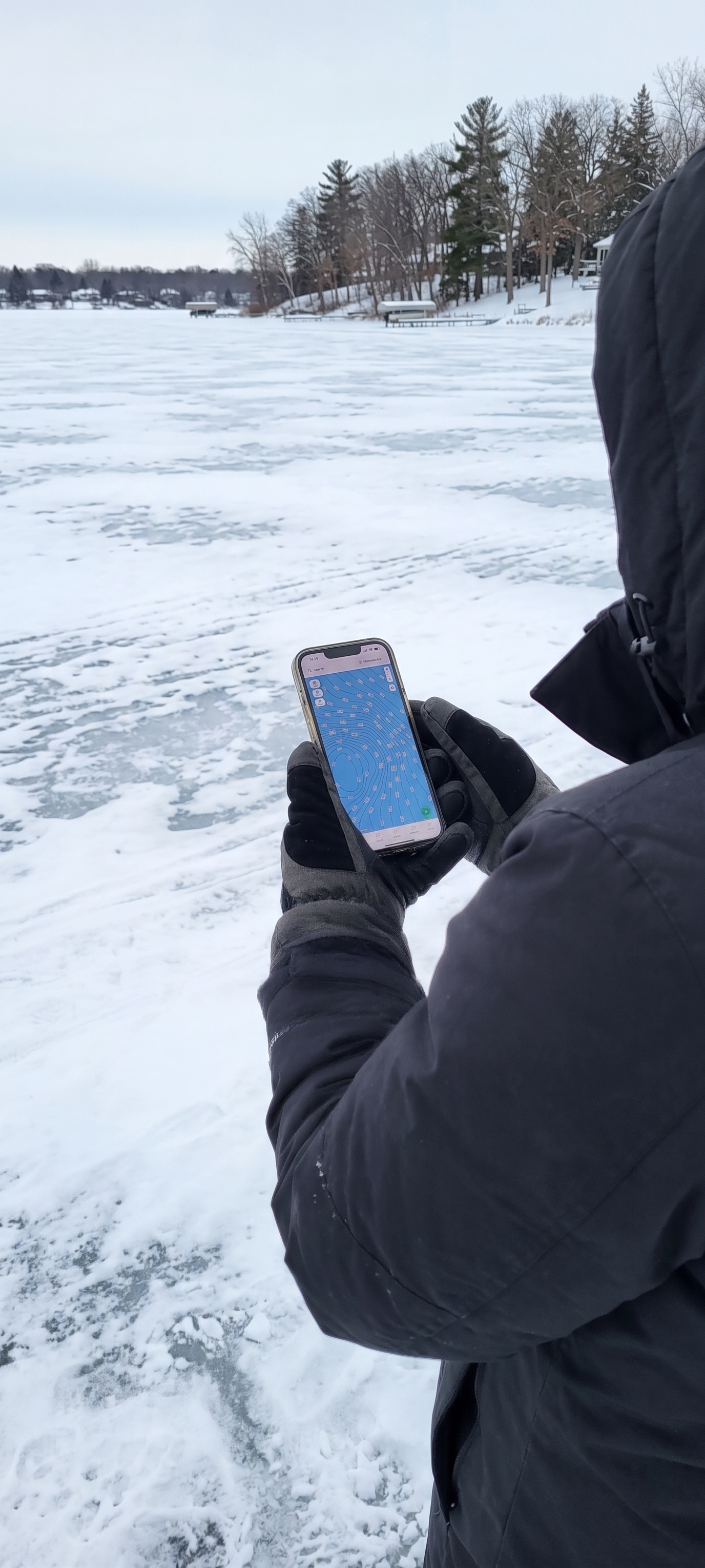An angler in a dark winter coat and gloves looks down at a smartphone showing a blue contour map of a Minnesota lake with a green waypoint pin, standing on patchy snow and bare ice with docks and a wooded shoreline behind.